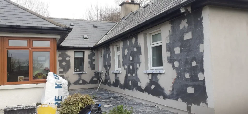 Exterior view of a house undergoing insulation installation, with insulation materials visible on the walls, before the siding is applied. Construction tools and equipment can be seen around the house, indicating ongoing work